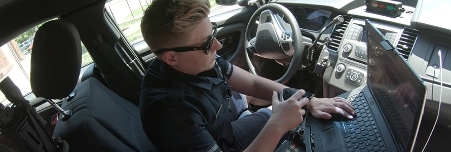 A police officer holds a radio and works on a mobile data computer while sitting in a patrol car.