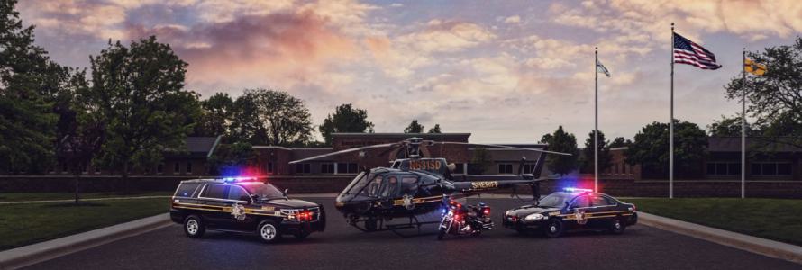 An Oakland County Sheriff’s Office SUV, helicopter, motorcycle, and cruiser with emergency lights activated sit outside at sunset