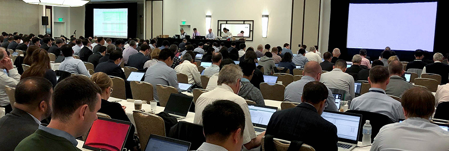 A room full of attendees at the June 2019 3GPP meetings listen and take notes on laptops as a panel of four speakers at the front give a presentation.