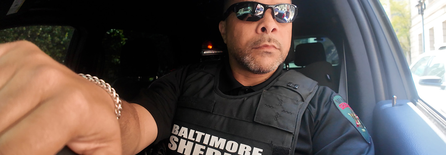 A Baltimore sheriff’s deputy driving a patrol vehicle with one hand on the steering wheel