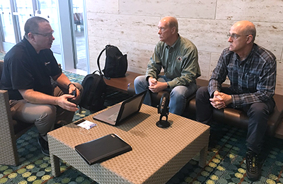 Brent Williams, Dr. Paul Zeeb, and Jack Rupp sit around a table which holds a laptop and a microphone.