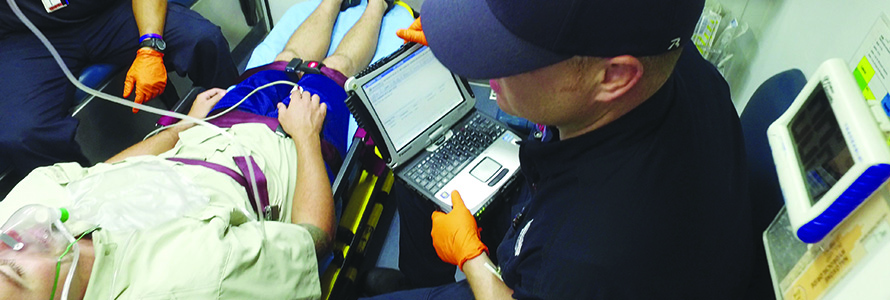 An EMS technician enters patient information into a laptop while riding in the back of an ambulance with a person in a stretcher