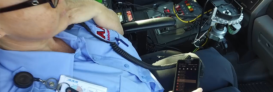 An emergency medical practitioner sits in an ambulance, speaking into a radio handset connected to a cell phone.