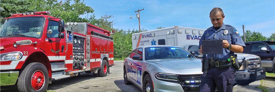 A police officer using a tablet stands in front of a fire truck, police car and ambulance.