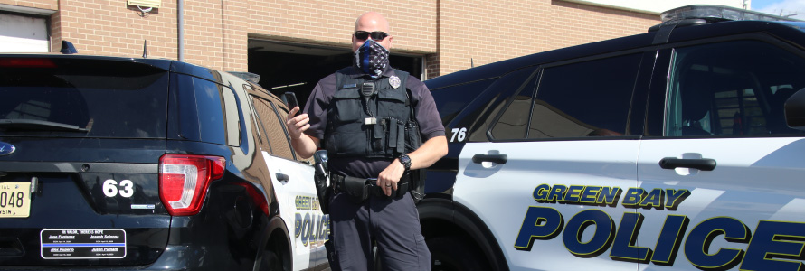 A police officer wearing a mask and a vest stands in front of a Green Bay Police vehicle