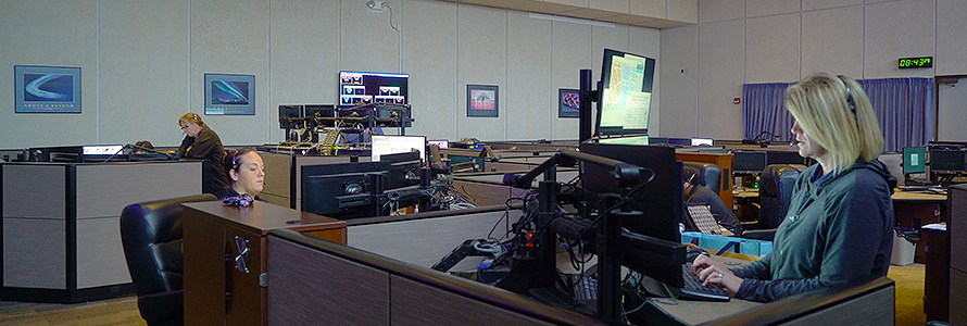 Three 9-1-1 call takers sit at dispatching consoles in a large 9-1-1 call center.