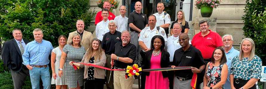 Group of officials and first responders standing in Allegany County, Maryland. Three people are holding a ceremonial ribbon, and one man cuts it with a large pair of scissors.