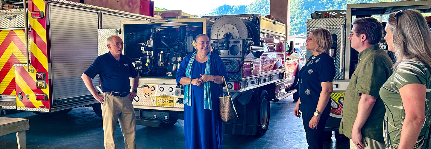 The FirstNet Authority’s Alexandra Fernández Navarro and Michael Varney and others talk with an official from the U.S. Pacific territories, standing at the rear of three public safety vehicles in an open apparatus bay.  