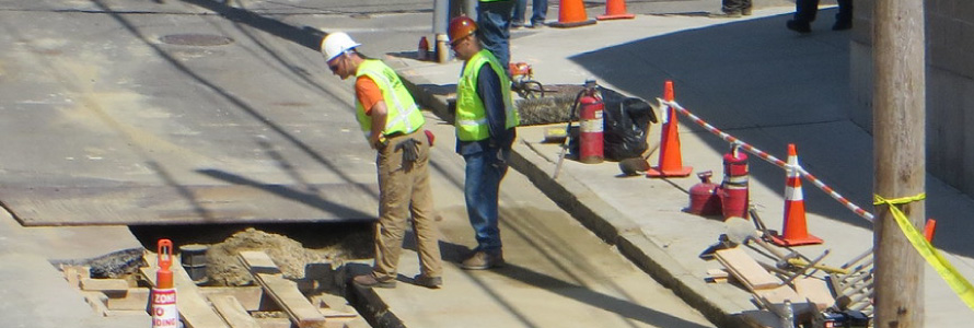 Workers in safety vests and hard hats observe a hole in the street.