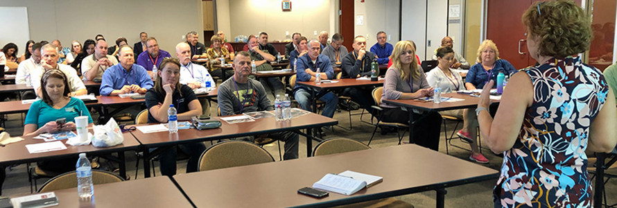 FirstNet Authority Senior Public Safety Advisor Kristi Wilde speaks in front of a group of emergency communications professionals seated behind desks 