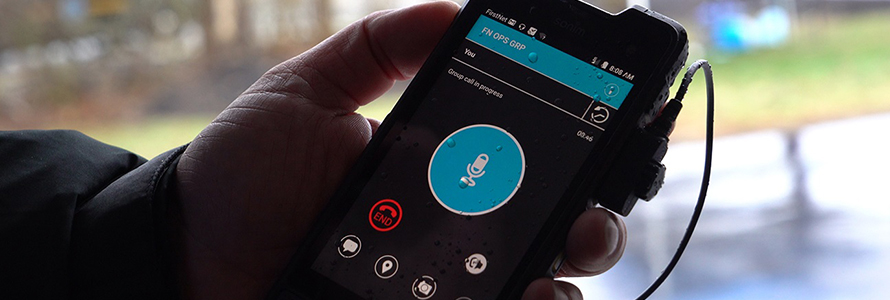 Hand holds FirstNet smartphone device with push-to-talk app displayed.