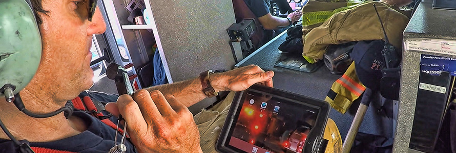 A firefighter riding in a helicopter speaks into a headset while looking down at a tablet with information