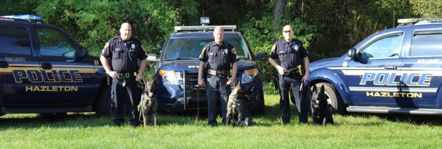 Three police officers standing in front of three Hazelton Police Department vehicles, each holding the leash of a German shepherd dog. 