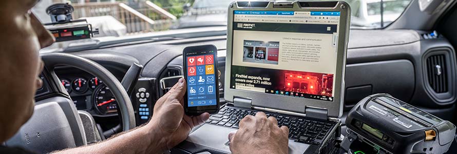 A police officer uses a FirstNet phone and a mobile computing device in his vhicle 