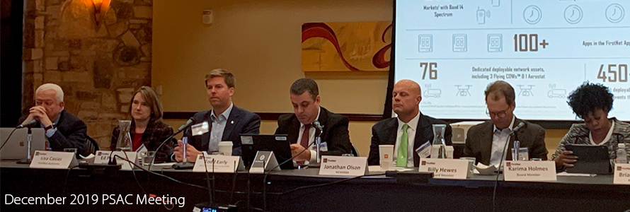 Members of the FirstNet Authority and the Public Safety Advisory Committee sit at table with slides projected behind; “December 2019 PSAC Meeting”