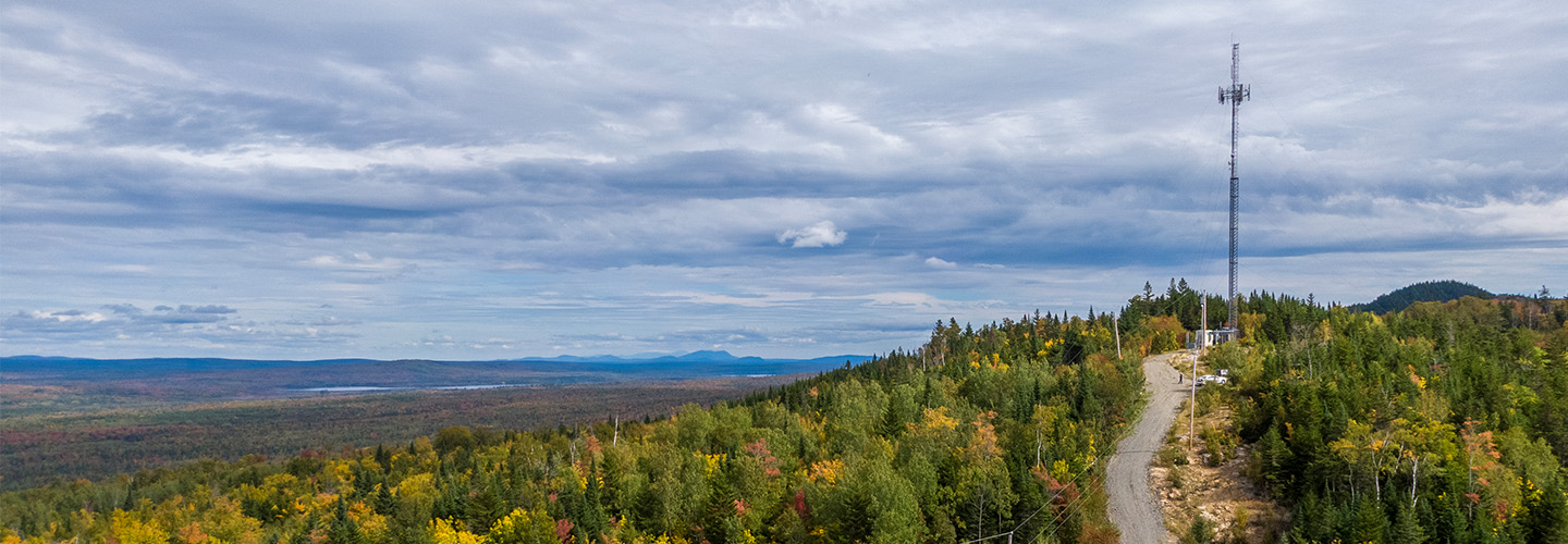 A FirstNet cell site situated on a hilly expanse surrounded by dense forest and a view of mountains and a lake in the distance.