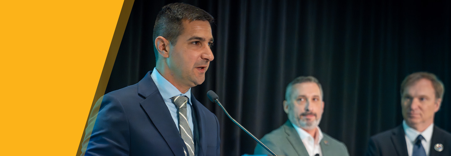 Chief Peter Burke of the Hyannis Fire Department in Massachusetts stands behind podium giving acceptance speech for the inaugural FirstNet Authority Patriot Award; FirstNet Authority Chief Market Engagement Officer Jeremy Zollo and Executive Director / CEO Joe Wassel look on