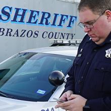 Deputy from Brazos Sheriff's Department looks at a smartphone standing in front of a mobile command vehicle