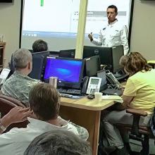 Emergency Manager addresses group of individuals including military uniformed men gesturing urgently.   