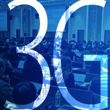 A group of people dressed in business attire taking notes on electronic devices while seated in a large conference room with three oversized presentation screens at the 3GPP 6G workshop; “3GPP”   
