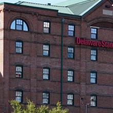 A blue light emergency phone sits to the right of a several story, campus building with Delaware State University across the facade. 