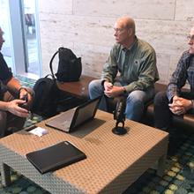 Brent Williams, Dr. Paul Zeeb, and Jack Rupp sit around a table which holds a laptop and a microphone.