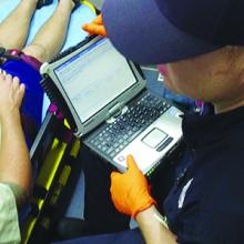 An EMS technician enters patient information into a laptop while riding in the back of an ambulance with a person in a stretcher