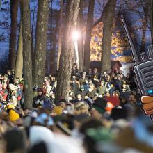A group of people gathered in a wooded area attending the annual Groundhog Day ceremony. A cartoon image of groundhogs dressed as emergency management officials with one holding a mobile device.