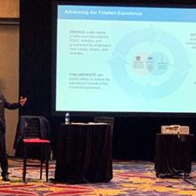 A man in a business suit stands in front of a presentation screen with his arm outstretched. 