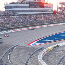 Race cars driving on the Iowa Speedway’s seven lane, oval racetrack in Newton, Iowa with thousands of spectators sitting in the surrounding stadium.