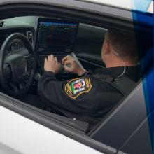 An officer in a Loudoun County Sheriff’s Office uniform sits at a steering wheel of a police vehicle using a mobile data computer mounted to the center console.  