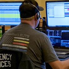 Emergency telecommunicators take calls at a desk with multiple monitors.