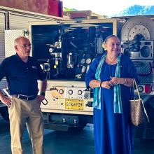 The FirstNet Authority’s Alexandra Fernández Navarro and Michael Varney and others talk with an official from the U.S. Pacific territories, standing at the rear of three public safety vehicles in an open apparatus bay.  