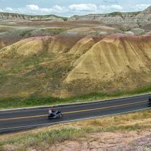 A scenic view of a winding road cutting through rocky hills in South Dakota, with a group of motorcyclists riding one behind the other along the curve of the road, surrounded by hills and rural terrain under a partly cloudy sky. A scenic view of a winding road cutting through rocky hills in South Dakota, with a group of motorcyclists riding one behind the other along the curve of the road, surrounded by hills and rural terrain under a partly cloudy sky.