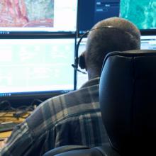 Swain County strengthens emergency communications with FirstNet 911 call-taker wearing a headset sitting at a desk in a communications center; multiple computer monitors showing maps, data, and emergency calls