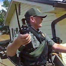 A first responder in tactical gear and holding a radio uses a pole and rope to assist a person through severe floodwaters outside a damaged home; Debris and muddy water surround the homes 