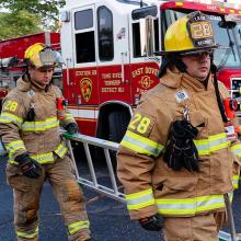 Two firefighters from the Toms River Fire Department in New Jersey carry a ladder while walking on a street in front of a fire truck 