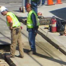 Workers in safety vests and hard hats observe a hole in the street. Workers in safety vests and hard hats observe a hole in the street.