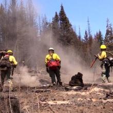 Smoke rises near five wildland firefighters working to put out remnants of a fire in an open space amongst trees.