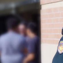 A female Sheriff’s deputy stands next to a brick wall and watches over a group of students.