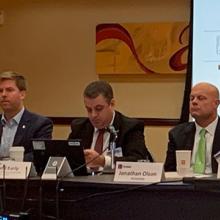 Members of the FirstNet Authority and the Public Safety Advisory Committee sit at table with slides projected behind; “December 2019 PSAC Meeting” Members of the FirstNet Authority and the Public Safety Advisory Committee sit at table with slides projected behind; “December 2019 PSAC Meeting”