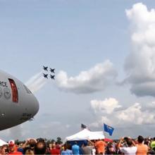 Four fighter jets streak across the sky on a summer day. In the foreground is a big crowd and a large aircraft.