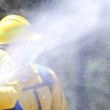 A first responder in a hard hat holds a hose spraying water; a second first responder in a hard hat stands behind.