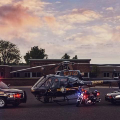 An Oakland County Sheriff’s Office SUV, helicopter, motorcycle, and cruiser with emergency lights activated sit outside at sunset