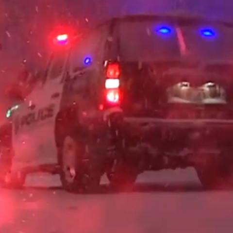 A police SUV with lights on and lit flare sit in a snowstorm at night.