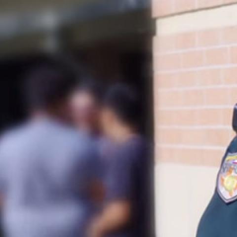 A female Sheriff’s deputy stands next to a brick wall and watches over a group of students.