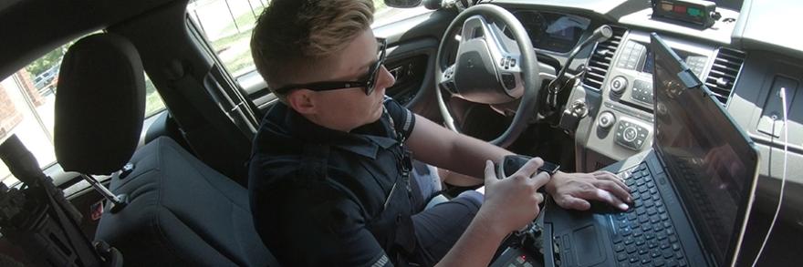 A police officer holds a radio and works on a mobile data computer while sitting in a patrol car.