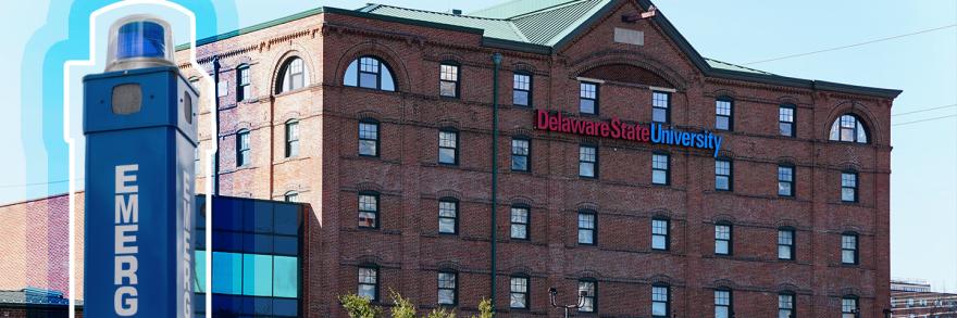A blue light emergency phone sits to the right of a several story, campus building with Delaware State University across the facade. 