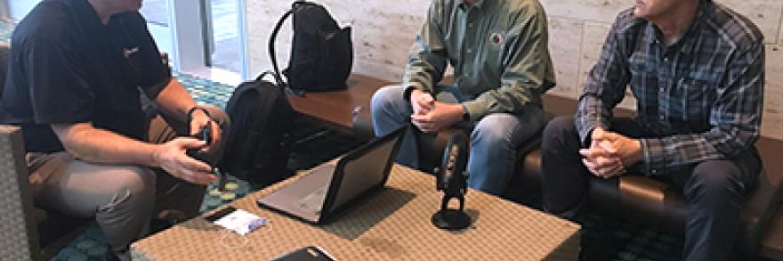 Brent Williams, Dr. Paul Zeeb, and Jack Rupp sit around a table which holds a laptop and a microphone.