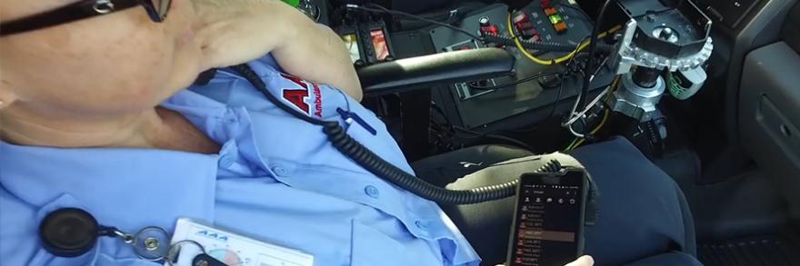 An emergency medical practitioner sits in an ambulance, speaking into a radio handset connected to a cell phone.